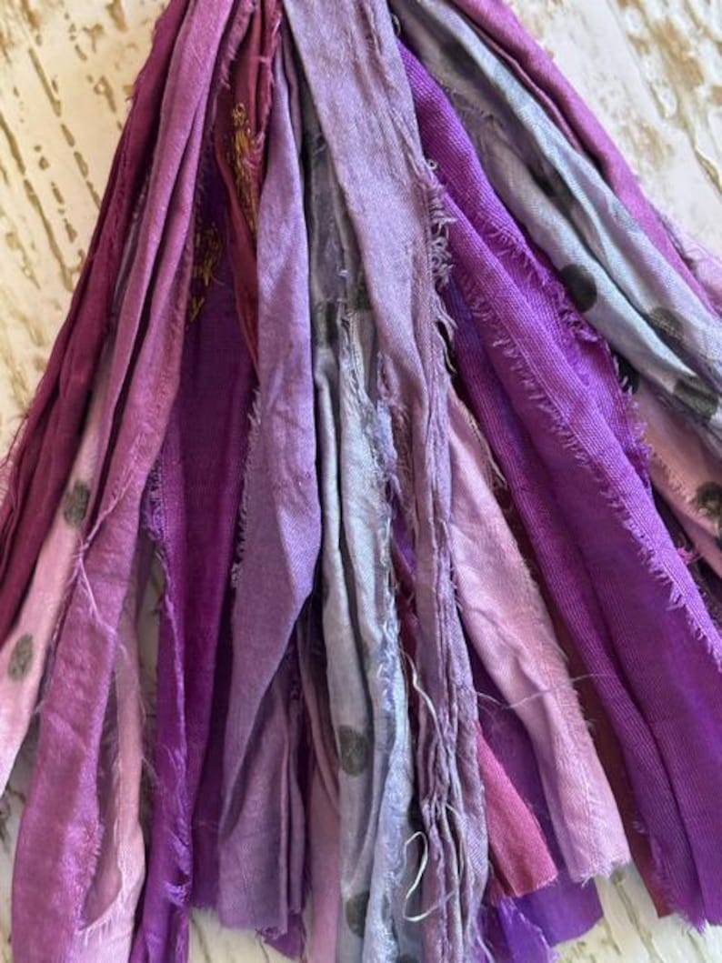 May include: A collection of fabric strips in various shades of purple, lavender, and light gray. The strips feature frayed edges and a mix of solid colors and patterns, including some with small dark polka dots, hanging vertically in a dense cluster.