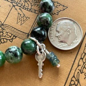 May include: A green and black gemstone bracelet with a silver key charm and a silver bead. The bracelet is on a brown book with the letters "FW" visible.