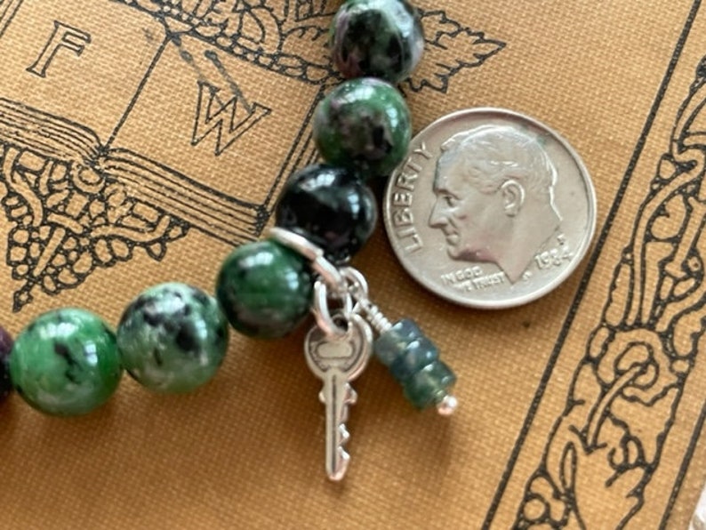 May include: A green and black gemstone bracelet with a silver key charm and a silver bead. The bracelet is on a brown book with the letters "FW" visible.