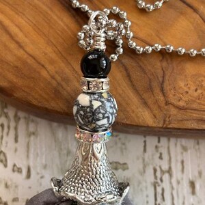 Puede incluir: Un collar plateado con un colgante decorativo. El colgante presenta una cuenta negra, una cuenta moteada y una base plateada texturizada. Una borla de tela gris cuelga de la base. El collar est&aacute; sobre una superficie de madera.
