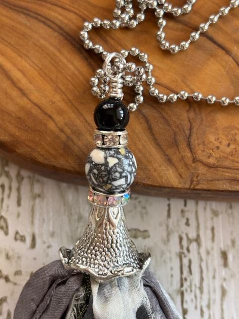 Puede incluir: Un collar plateado con un colgante decorativo. El colgante presenta una cuenta negra, una cuenta moteada y una base plateada texturizada. Una borla de tela gris cuelga de la base. El collar est&aacute; sobre una superficie de madera.