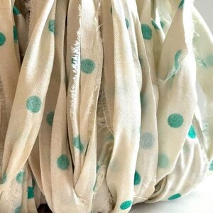 May include: Close-up of a bundle of off-white fabric strips with teal polka dots. The fabric has a slightly frayed edge, and the strips are arranged in a layered, textured pattern. The overall effect is soft and visually appealing.