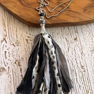 Puede incluir: Un collar con una cadena de bolas plateada y una borla grande hecha de tiras de tela superpuestas en tonos negro, gris y blanco. La borla est&aacute; asegurada con una tapa de metal plateado y una cuenta con un patr&oacute;n en blanco y negro.