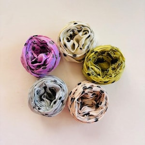 Puede incluir: Seis pañuelos de seda enrollados en forma circular. Los pañuelos son de varios colores, incluyendo lavanda, crema, amarillo verdoso, gris claro y melocotón. Cada pañuelo tiene un estampado de pequeños lunares negros.