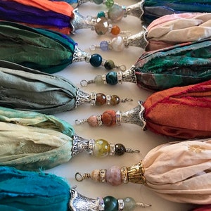 May include: A collection of decorative tassels made from strips of colorful silk fabric. Each tassel features a silver-toned metal cap adorned with various gemstone beads and small crystal accents, ending in a wire loop for attachment.