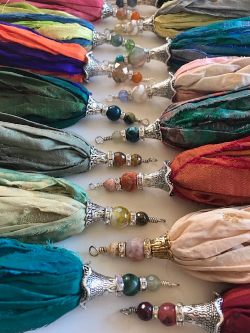 May include: A collection of decorative tassels made from strips of colorful silk fabric. Each tassel features a silver-toned metal cap adorned with various gemstone beads and small crystal accents, ending in a wire loop for attachment.