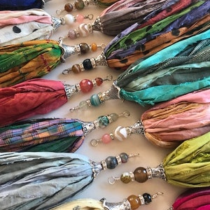 May include: A collection of decorative tassels made from various colorful silk ribbons. Each tassel features a silver-toned metal cap and a series of multi-colored gemstone beads attached to a wire loop at the top for hanging or attachment.