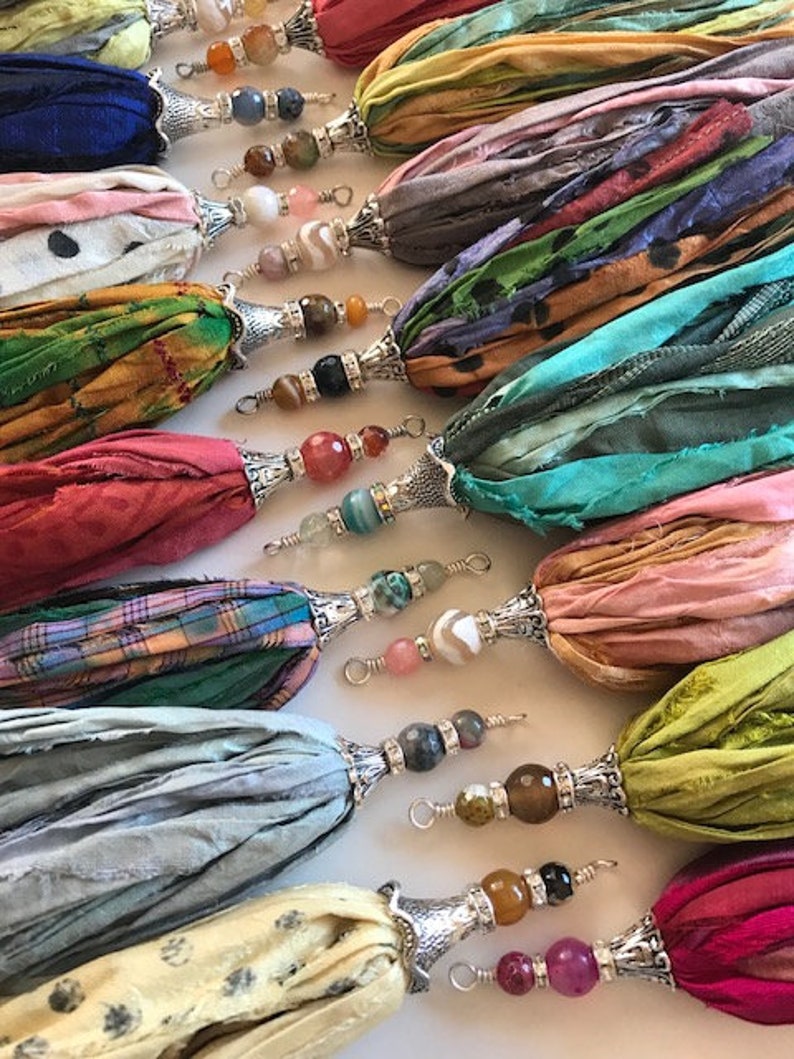 May include: A collection of decorative tassels made from various colorful silk ribbons. Each tassel features a silver-toned metal cap and a series of multi-colored gemstone beads attached to a wire loop at the top for hanging or attachment.