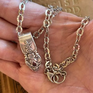 May include: A silver-toned necklace featuring a pendant crafted from the handle of a vintage spoon with an embossed floral pattern. The pendant is attached to a textured oval link chain that secures with a lobster claw clasp closure.