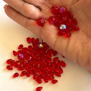 May include: A collection of small, translucent red beads in various shapes, including round faceted spheres and teardrop forms. Some of the round beads feature an iridescent finish that reflects light, while the teardrop beads are smooth and solid red.