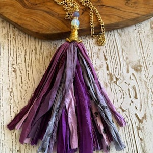 May include: A necklace featuring a gold-tone chain and a large tassel made of multiple strips of fabric in various shades of purple and lavender. The tassel is topped with a gold-tone metal cap, a small white bead, and a small purple bead.