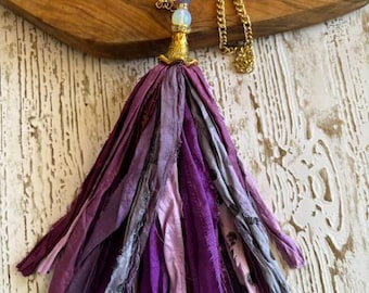 Puede incluir: Un collar con una cadena de tono dorado y una borla grande hecha de múltiples tiras de tela en varios tonos de morado y lavanda. La borla está rematada con una tapa de metal dorado, una cuenta blanca pequeña y una cuenta morada.