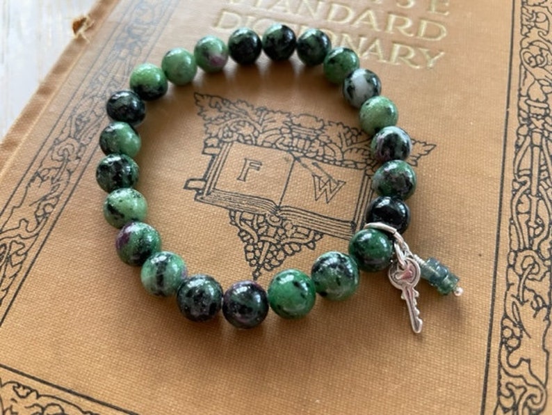 May include: A green and black gemstone bracelet with a silver key charm. The bracelet is made of round beads and is lying on a book.