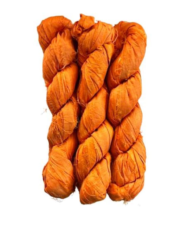 May include: A bundle of three twisted skeins of vibrant orange silk ribbon yarn. The material features a crinkled, textured surface with frayed edges along the length of the fabric strips, creating a voluminous and layered appearance in this yarn.