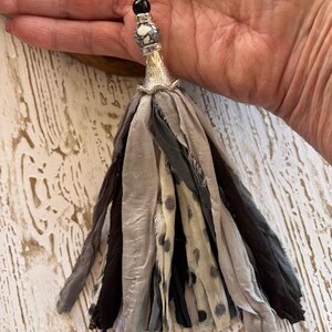 Puede incluir: Un collar con colgante de borla hecho a mano con una cadena plateada. La borla est&aacute; hecha de tiras de tela en capas en tonos de gris, negro y crema con lunares negros. El colgante presenta un cono plateado, una cuenta estampada y una cuenta negra.