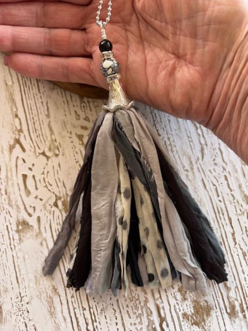Puede incluir: Un collar con colgante de borla hecho a mano con una cadena plateada. La borla est&aacute; hecha de tiras de tela en capas en tonos de gris, negro y crema con lunares negros. El colgante presenta un cono plateado, una cuenta estampada y una cuenta negra.