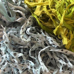 May include: A close-up of fabric strips in blue, green, and yellow. The blue strips have a polka dot pattern. The green and yellow strips are solid colours. The fabric appears to be silk or a similar material. Ideal for craft projects.