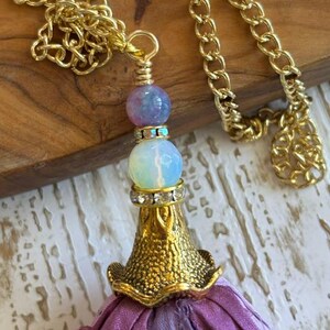 May include: A gold-tone chain necklace featuring a pendant with a purple marbled bead, a translucent white bead, and a gold-tone metal cap with clear rhinestones. A long, frayed purple and grey fabric tassel hangs from the metal cap.