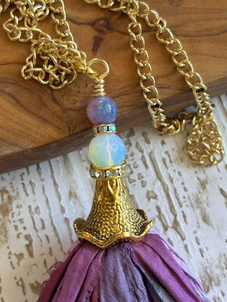 May include: A gold-tone chain necklace featuring a pendant with a purple marbled bead, a translucent white bead, and a gold-tone metal cap with clear rhinestones. A long, frayed purple and grey fabric tassel hangs from the metal cap.