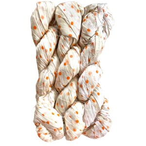 May include: A skein of off-white silk ribbon yarn featuring a repeating pattern of small, vibrant orange polka dots. The ribbon has a crinkled, textured appearance and is twisted into a bundle, showcasing the soft sheen of the fabric material.