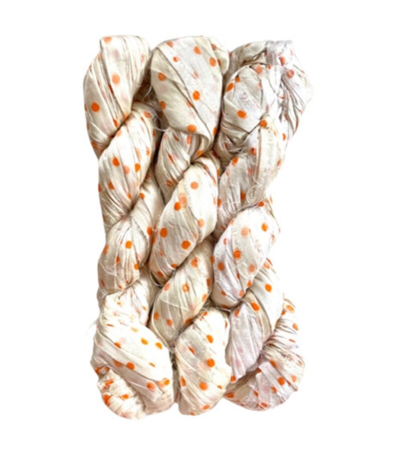 May include: A skein of off-white silk ribbon yarn featuring a repeating pattern of small, vibrant orange polka dots. The ribbon has a crinkled, textured appearance and is twisted into a bundle, showcasing the soft sheen of the fabric material.