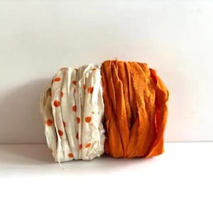 May include: Two bundles of fabric ribbon are positioned side by side. One bundle is off-white with small orange polka dots, while the other bundle is a solid, vibrant orange. Both ribbons feature frayed, raw edges and a textured, crinkled appearance.