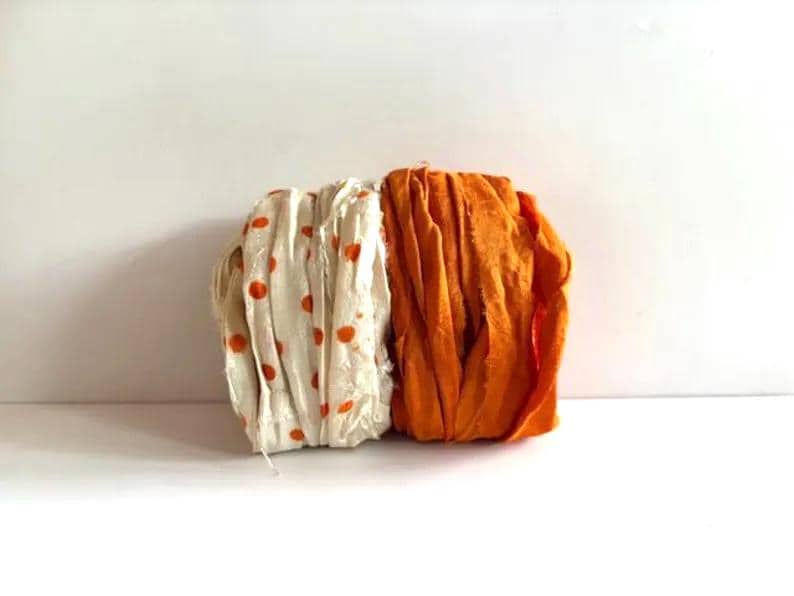 May include: Two bundles of fabric ribbon are positioned side by side. One bundle is off-white with small orange polka dots, while the other bundle is a solid, vibrant orange. Both ribbons feature frayed, raw edges and a textured, crinkled appearance.