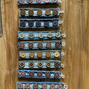 Könnte beinhalten: Zehn Armbänder aus Denim mit weißer Spitze und bunten, gemusterten Knöpfen. Jedes Armband hat ein anderes Knopfdesign.