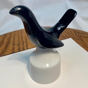 May include: A glossy black ceramic bird figurine perched atop a smooth, cylindrical white base. The bird features a curved neck, a pointed beak, and an angled tail, creating a sleek silhouette. The entire piece has a reflective finish.