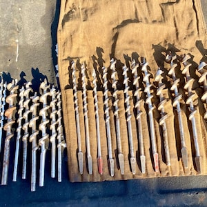 May include: A collection of silver-colored metal auger drill bits of varying diameters, arranged in a row and alongside a brown canvas tool roll with individual slots. The drill bits feature a spiral flute design and tapered shanks.