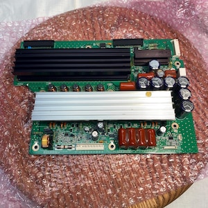 May include: A green circuit board with various electronic components, including a black heat sink, silver heat sink, and several capacitors. The board is on a wooden surface and wrapped in pink bubble wrap.
