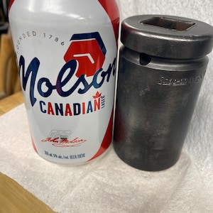 Pode incluir: Uma lata de cerveja Molson Canadian com detalhes em vermelho e branco, e um soquete de chave Snap-on preto. A lata apresenta o logotipo da Molson e o texto "CANADIAN LAGER". O soquete é cilíndrico.