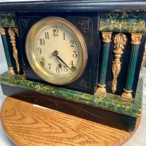 Puede incluir: Reloj de chimenea negro antiguo con esfera y manecillas doradas. El reloj presenta detalles dorados y verdes, incluyendo columnas decorativas y una cabeza de león. Se apoya sobre una base de madera.