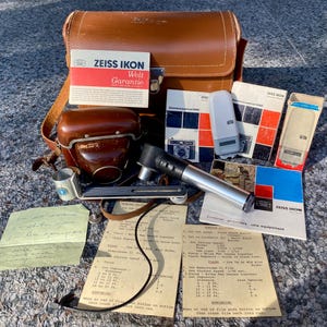 May include: A brown leather camera bag containing a Zeiss Ikon camera case, a silver and black flash unit, a metal film advance crank, and several printed manuals. Included are two beige instruction sheets with black text and a handwritten note.