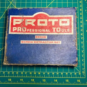 Op de afbeelding: Een blauwe doos met de tekst "PROTO PROFESSIONAL TOOLS" in rood en wit. De doos heeft ook de tekst "9500B SCREW EXTRACTOR SET". De doos staat op een groen oppervlak met inch- en centimetermarkeringen.