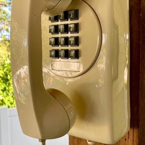 May include: A cream-coloured wall-mounted telephone with a matching handset resting on the cradle. The device features a black push-button keypad with numbers 0-9, an asterisk, and a hash sign. A coiled cord connects the handset to the base unit.