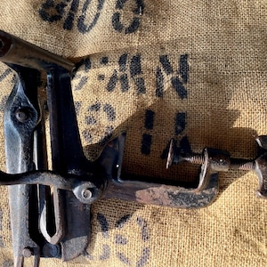May include: A vintage cast iron saw set tool featuring a dark, weathered metal finish. The device includes a mechanical clamping mechanism with a threaded screw handle, a curved lever, and a notched jaw assembly designed for adjusting saw teeth.