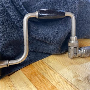 Puede incluir: Un taladro manual antiguo con un mango y pomo de madera negra. El taladro tiene un marco de metal plateado y un portabrocas para sujetar brocas. Descansa sobre una superficie de madera con un paño azul oscuro en el fondo.