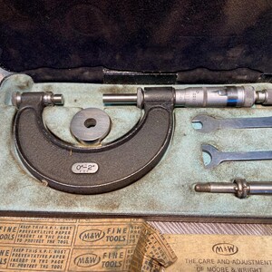 May include: A vintage black micrometer set with a 0-2 inch range, including small wrenches and a cylindrical piece, all nestled in a blue-green velvet-lined case. The case is open, revealing the tools. The set includes the text "M&W FINE TOOLS".
