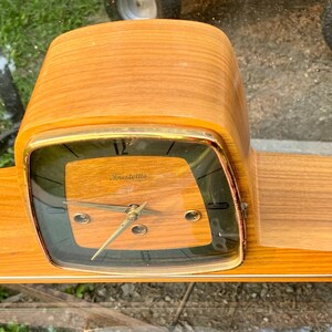 Puede incluir: Un reloj de sobremesa con acabado de veta de madera marrón claro y una base horizontal ancha. La esfera es cuadrada con esquinas redondeadas, con un marco metálico dorado, marcadores de hora negros y manecillas doradas finas tras un cristal.