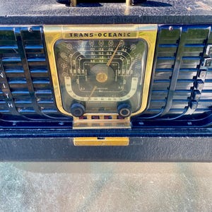 May include: A vintage Zenith Trans-Oceanic portable radio featuring a black textured case, a gold-toned central dial with frequency markings, and vertical black slats. The front panel displays the text Zenith, Trans-Oceanic, and Best Reception.