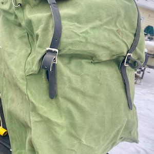 May include: A large, olive green canvas backpack with black leather straps and silver buckles. The top edge is trimmed with red fabric. The bag appears to be vintage or military-style, suitable for outdoor activities or travel.