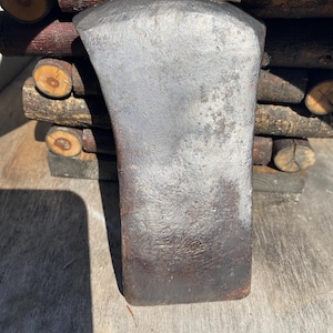 May include: A heavy, dark gray metal axe head with a wide, curved cutting edge and a weathered, textured surface. The metal shows signs of oxidation and wear, featuring a flat poll and a tapered body designed for attachment to a handle.
