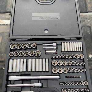 May include: A black plastic Craftsman tool case containing a comprehensive set of silver metal socket wrenches, various sized sockets, and three ratchets. The interior is molded to hold each tool securely in its designated labeled compartment.