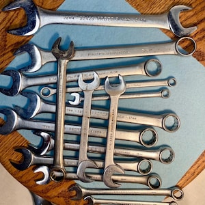 Puede incluir: Una colección de llaves inglesas plateadas de varios tamaños y formas, dispuestas sobre una superficie azul. Algunas llaves tienen texto como "PROTO" y marcas de medición. Herramientas para trabajos mecánicos o de reparación.