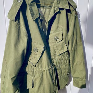 May include: An olive green field jacket featuring a collared neckline, long sleeves with buttoned cuffs, and four front pockets with buttoned flaps. The jacket includes shoulder epaulets, a drawstring waist, and a front button closure system.