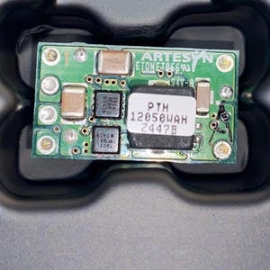 Puede incluir: Primer plano de una placa de circuito verde con varios componentes electrónicos. La placa tiene el texto "ARTESYN" y "PTH 12050UAH 2447B". La placa está en una bandeja negra.