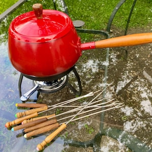 May include: A bright red fondue pot with a matching lid, featuring a wooden handle and a wooden lid knob. The pot rests on a black metal stand with a chrome burner. A set of six metal fondue forks with wooden handles and coloured tips is included.