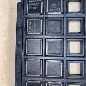 以下が含まれることがあります： Xilinx XCR3064XL集積回路が複数入った黒いトレイ。各チップは濃いグレーで、Xilinxのロゴやモデル情報などの白い文字が記載されています。トレイには正方形のコンパートメントがあります。