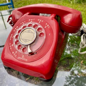 May include: A bright red rotary telephone featuring a circular dial with numbers and letters, a central blank label, and a matching handset resting on top. The device includes a coiled cord extending from the side and a classic desk-style base.